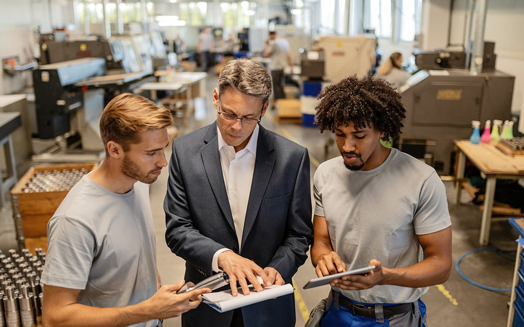 Factory manager discussing quality control reports with two young engineers using digital tablets in an industrial workshop.